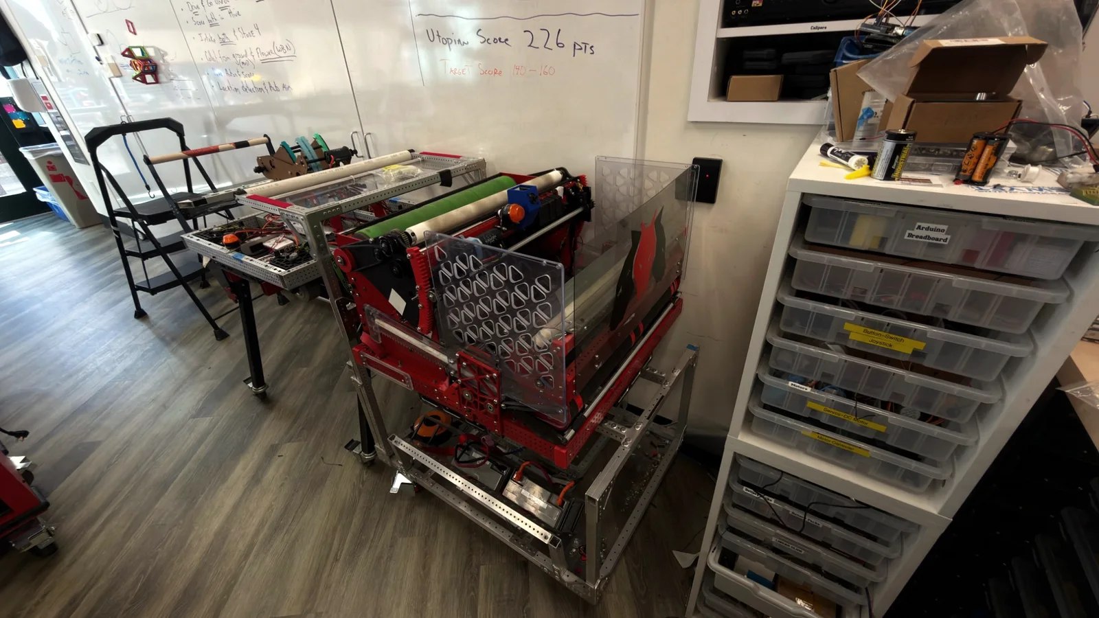 Team 2584’s robot on its cart in the workshop: a red frame with green and white intake rollers and clear side panels, in front of a whiteboard listing the season’s scoring goals